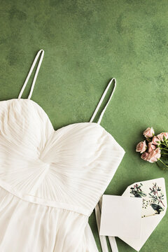 Studio shot of white wedding dress, invitation card and roses flat laid against green background