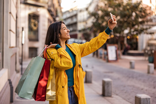 Happy woman with shopping bags taking selfie over smart phone on footpath