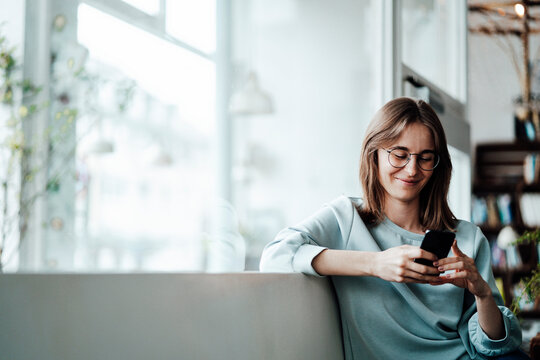 Smiling woman text messaging through smart phone sitting in cafe