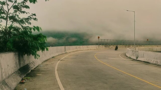 A winding road disappears into thick, atmospheric mist blanketing distant mountains, creating a serene and solitary journey through a mystical, fog-laden landscape