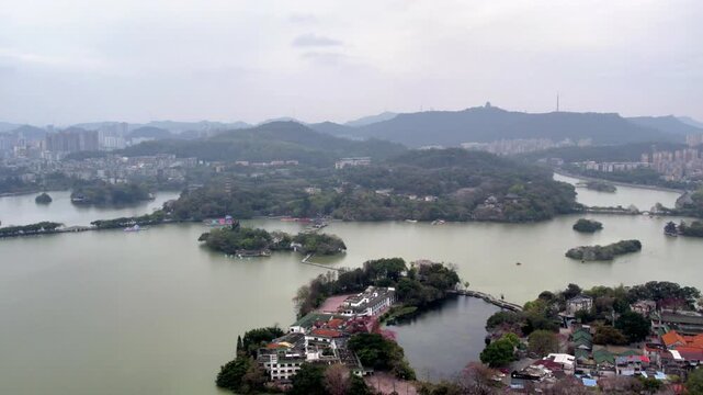 This is an elevated view of a lake with multiple tree-covered islands, bordered by urban areas and rolling hills under an overcast sky.
