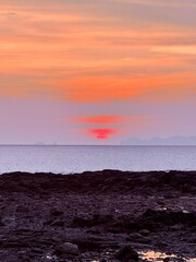 Obraz premium Stunning tropical sunset with a glowing red sun sinking below the horizon, soft pastel clouds, calm ocean, and rocky shoreline creating a peaceful coastal landscape.
