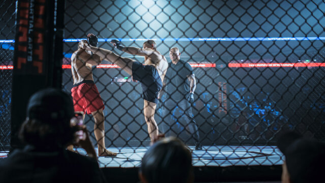 Two Fighters Clash in an MMA Fight as One Lands a High Kick to the Opponent&rsquo;s Head Inside the Cage. a Referee Tracks the Action While Fans Film Through the Fence, Fight for the Champion Title Belt.