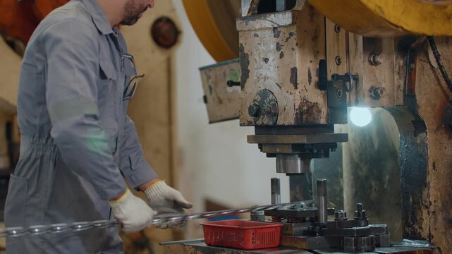 Worker in factory wears hard hat and gloves. He handles metal parts with focused, concentrated expression. Large machine dominates foreground with industrial machinery behind