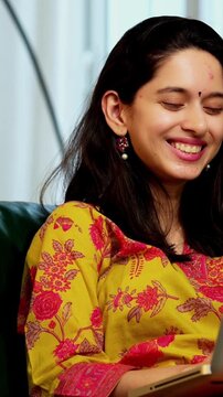 Confident Indian Asian young woman in her mid-twenties wearing a kurta, sitting on a sofa in a cozy living space, video chatting on a laptop using wireless headphones