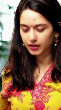 Confident Indian Asian young woman in her mid-twenties wearing a kurta, sitting on a sofa in a cozy living space, video chatting on a laptop using wireless headphones