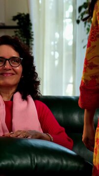 Closeup portrait of a happy Indian Asian pretty young girl in her twenties and her mother in fifties, both looking at the camera while sitting close together on a cozy sofa at home, enjoying their tim