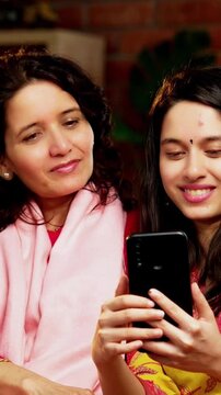 Indian Asian pretty young girl in her twenties and her mother in fifties sitting together on a sofa at home, both using a smartphone, enjoying cozy time and connection in a comfortable living room