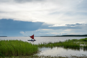 Small sailing boat with a distinctive red sail on calm coastal waters under dramatic summer clouds in Österby, Läänemaa, Estonia. © ago
