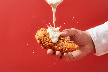 Hand Holding Crispy Chicken Tenders with Ranch Sauce Pouring on Red Background A hand holds crispy golden fried chicken tenders as creamy ranch sauce pours and splashes on top, captured in a bold red  © kamal ud din