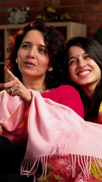 Indian Asian mother in her fifties with her pretty young daughter, both sitting on a cozy sofa at home, watching TV together with a remote in hand