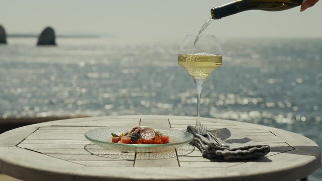 White wine being poured into glass with seafood on wooden table by ocean