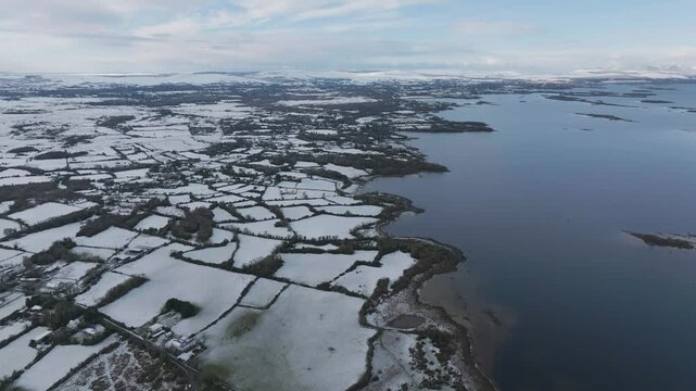 A cinematic aerial shot showcasing the snowcovered rural landscape and coastline of Lough Corrib, Ireland, featuring patchwork fields and calm winter waters.