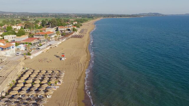 Aerial tracking shot of Kourouta beach bars lounge areas, sunbeds and natural straw umbrellas in the shallow waters of Ionian Sea, Ilia