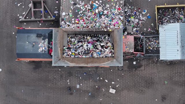 Plastic waste is loaded into a large container for sorting and processing. Workers separate recyclable materials from non-recyclable waste. The facility operates in an industrial area.
