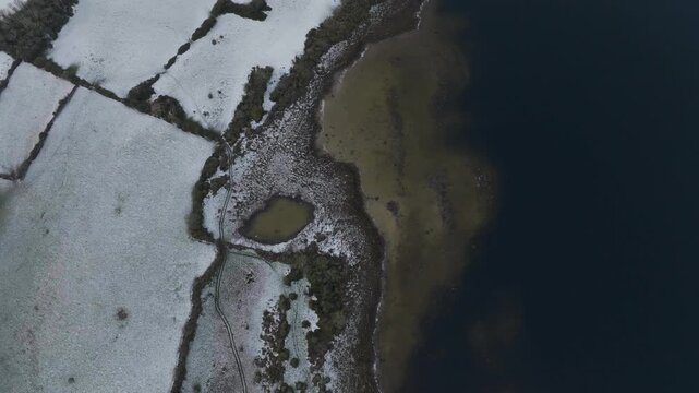 A cinematic topdown aerial shot showing the contrast between snowcovered Irish fields and the dark waters of Lough Corrib during winter.