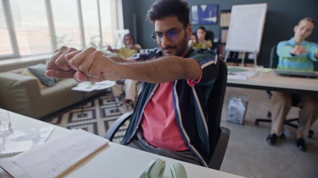 Stab shot of young adult Indian man stretching hands and arms at desk with multiethnic colleagues in background. Clip is perfect for corporate wellness programs and office ergonomics training