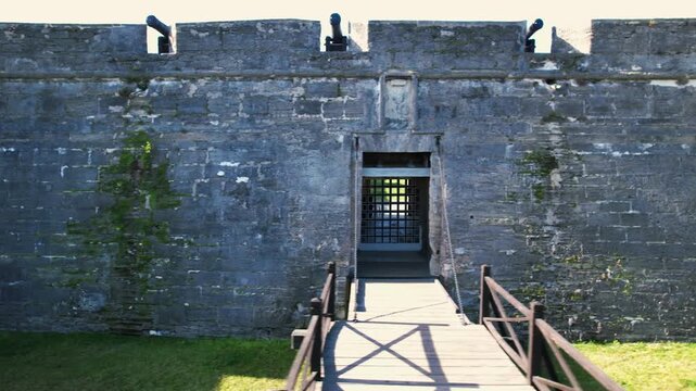 Ancient stone fortress drawbridge access with cannons on parapet overlooking green grass.