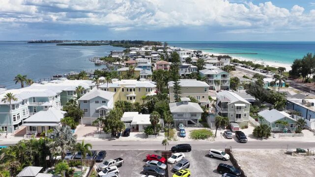 Colorful beach houses line coastal street, turquoise Gulf waters, sandy beach, palm trees, parked cars, summer clouds, distant island, Bradenton, Florida, United States. Aerial drone wide shot