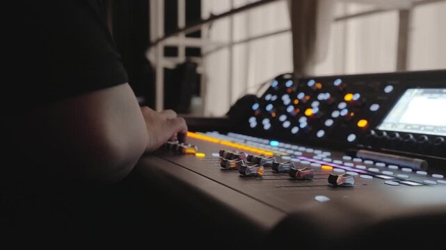 Close-up of a hand touching faders on a black digital mixing console with orange glowing lights, knobs, and a display screen at a live event venue.