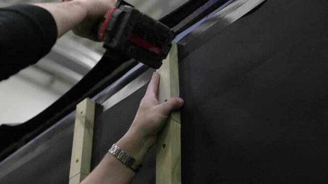 Worker fastening vertical wood framing to a dark wall panel with a cordless drill during house construction