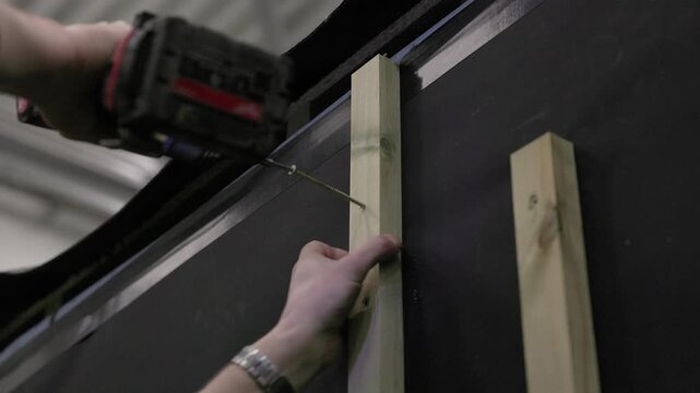 Worker fastening vertical wood framing to a dark wall panel with a cordless drill during house construction