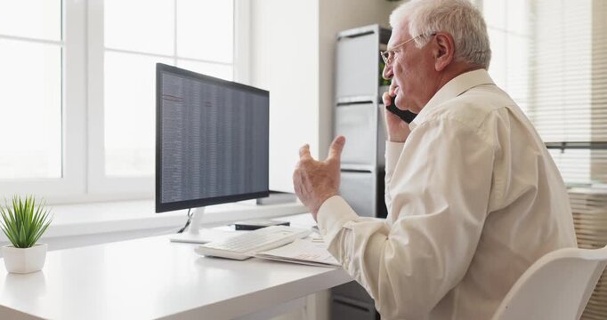 Senior man accountant on phone call. In a bright office analyst reviews spreadsheets on a computer discussing data and analysis with a client. Concept is finance and business communication.