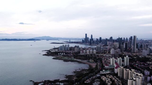 A panoramic aerial view of a coastal city skyline adjacent to calm sea, with distant land and overcast sky.