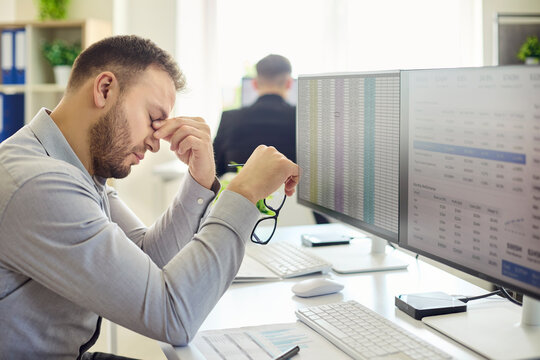 Employee man shows stress at computer spreadsheet. An office analyst rubs eyes at monitors, facing deadline errors and data work causing overwork and fatigue. Symbol of burnout at office.