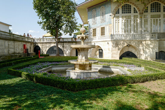 Classical multi-tiered fountain in the sunlit Topkapi Palace gardens, surrounded by a symmetrical lavender parterre and backed by the iconic blue-tiled Ottoman pavilion.