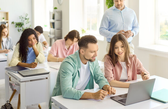 Students in classroom learning during lesson with teacher. Two young adults collaborate at a laptop while the instructor supervises progress. Teamwork and collaborative study in college settings.