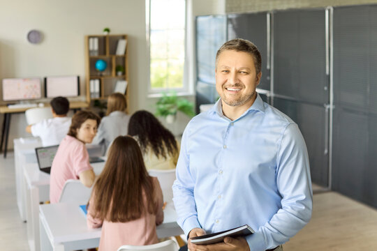Teacher students classroom lesson learning education. Smiling male professor stands by desks while students work on laptops in a bright modern classroom. Clear teaching and leadership values.