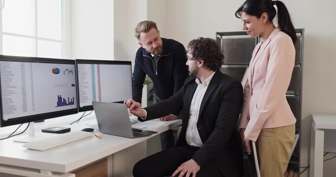 Team colleagues meeting in office for analysis. They review data and charts on dual monitors while a manager points to key insights on a laptop. Teamwork and collaboration drive results for business.