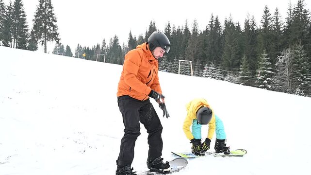 Two snowboarders prepare on a snow-covered slope. One assists the other. Trees frame the background in a winter wonderland setting.