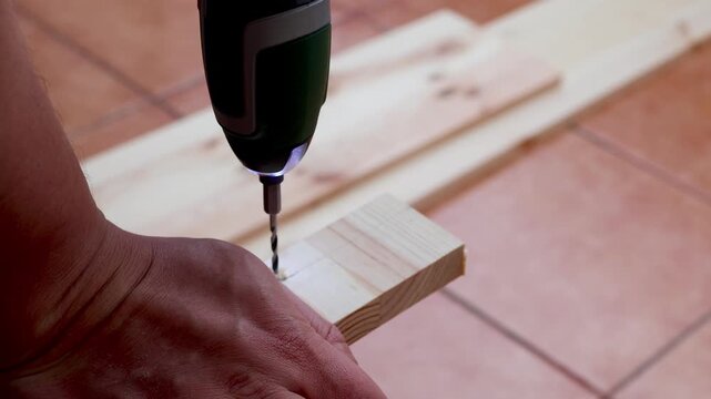 Man drills a pilot hole into a wooden board with a cordless drill during a DIY furniture project on balcony. Concept of woodworking, home improvement, furniture assembly and practical building skill.