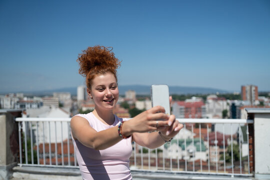 Beautiful and young redhead woman taking a selfie with a mobile phone on the rooftop of the city building