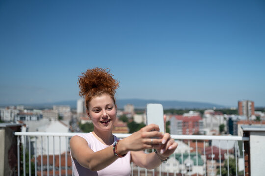 Beautiful and young redhead woman taking a selfie with a mobile phone on the rooftop of the city building