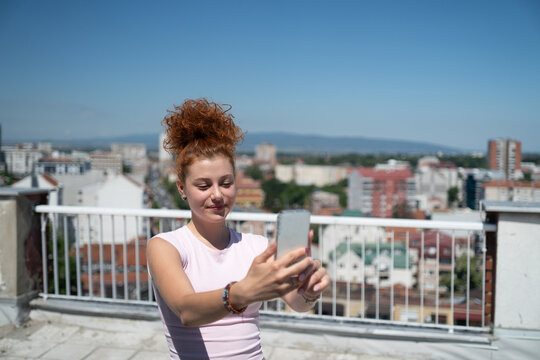 Beautiful and young redhead woman taking a selfie with a mobile phone on the rooftop of the city building
