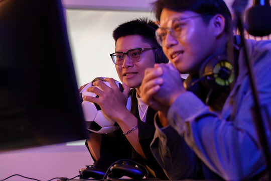 Two Asian men watching soccer e-sports on computer with football for FIFA World Cup anticipation