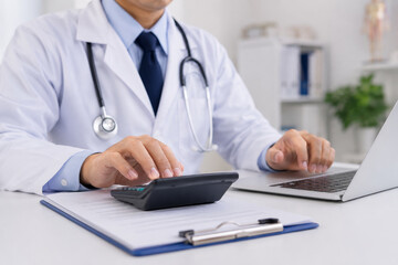 Fototapeta premium Close up of a medical professional calculating expenses. Doctor hands using a calculator and laptop for patient insurance claims, medical bills, and clinic budget planning.