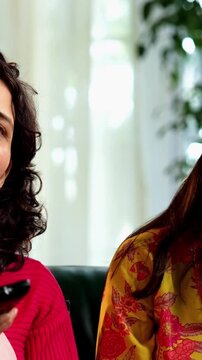 Indian Asian mother in her fifties with her pretty young daughter, both sitting on a cozy sofa at home, watching TV together with a remote in hand