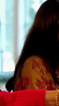 Indian Asian mother in her fifties with her pretty young daughter, both sitting on a cozy sofa at home, watching TV together with a remote in hand