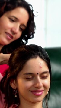 Indian Asian mother in her fifties lovingly giving an oil head massage to her pretty young daughter while sitting together on a sofa at home, expressing care and affection