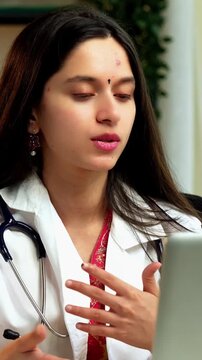 Indian Asian young female doctor in her mid-twenties, wearing a white coat, sitting at a desk with a laptop, conducting an online consultation with patients via video call in a professional setup