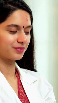 Indian Asian pretty young female dentist in her mid-twenties wearing a coat, seated at a desk, smiling confidently while looking directly at the camera in a professional setting