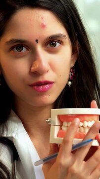 Indian Asian young female dentist in her mid-twenties wearing a coat, seated at a desk, smiling and explaining oral hygiene using a model denture while looking at the camera