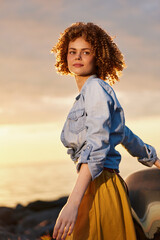 Naklejka premium A woman with curly hair stands beside a coastal road at sunset, wearing a denim shirt and mustard skirt, relaxed and thoughtful. The warm glow creates a calm lifestyle moment outdoors.