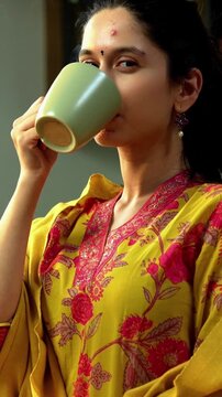 Indian Asian happy and pretty young girl in her twenties wearing salwar kameez and shawl, enjoying hot coffee or beverage in a modern kitchen with warm morning sunlight streaming in