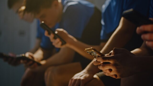Young Male Football Players Relaxing on Benches in Sports Changing Room, Silently Using Smartphones. Concept of Modern Youth Digital Routine, Digital Isolation Among Gen Z Athletes, Youth Lifestyle.