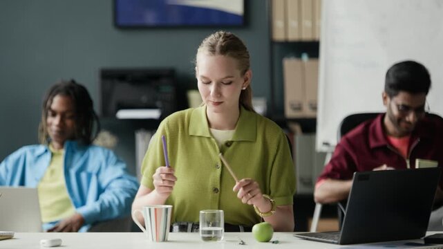 Waist up of Caucasian young adult woman tapping pen and pencil on desk items like drums in office, multiethnic colleagues working in background. Clip represents office distraction or creative block